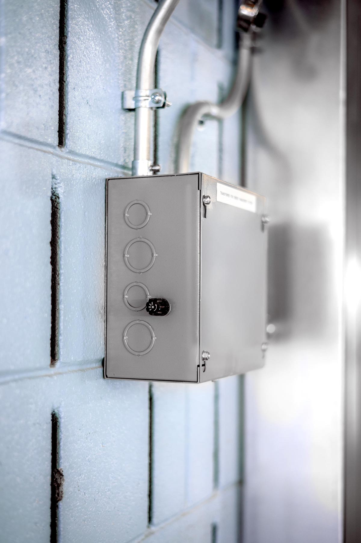 Close-up of a gray metal electrical box mounted on a light blue concrete block wall with conduits running above it.