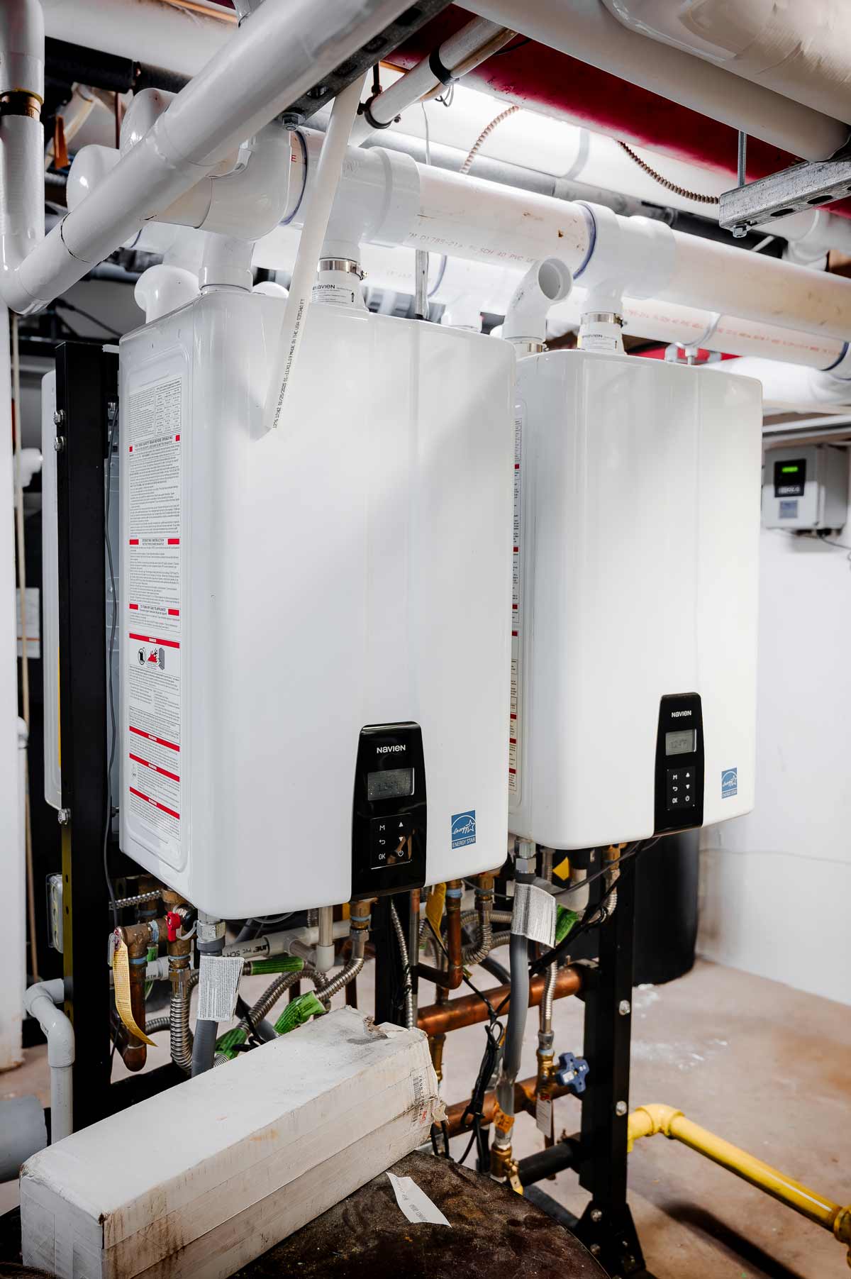 Two white Navien tankless water heaters installed side by side with visible plumbing and pipes in a utility room.