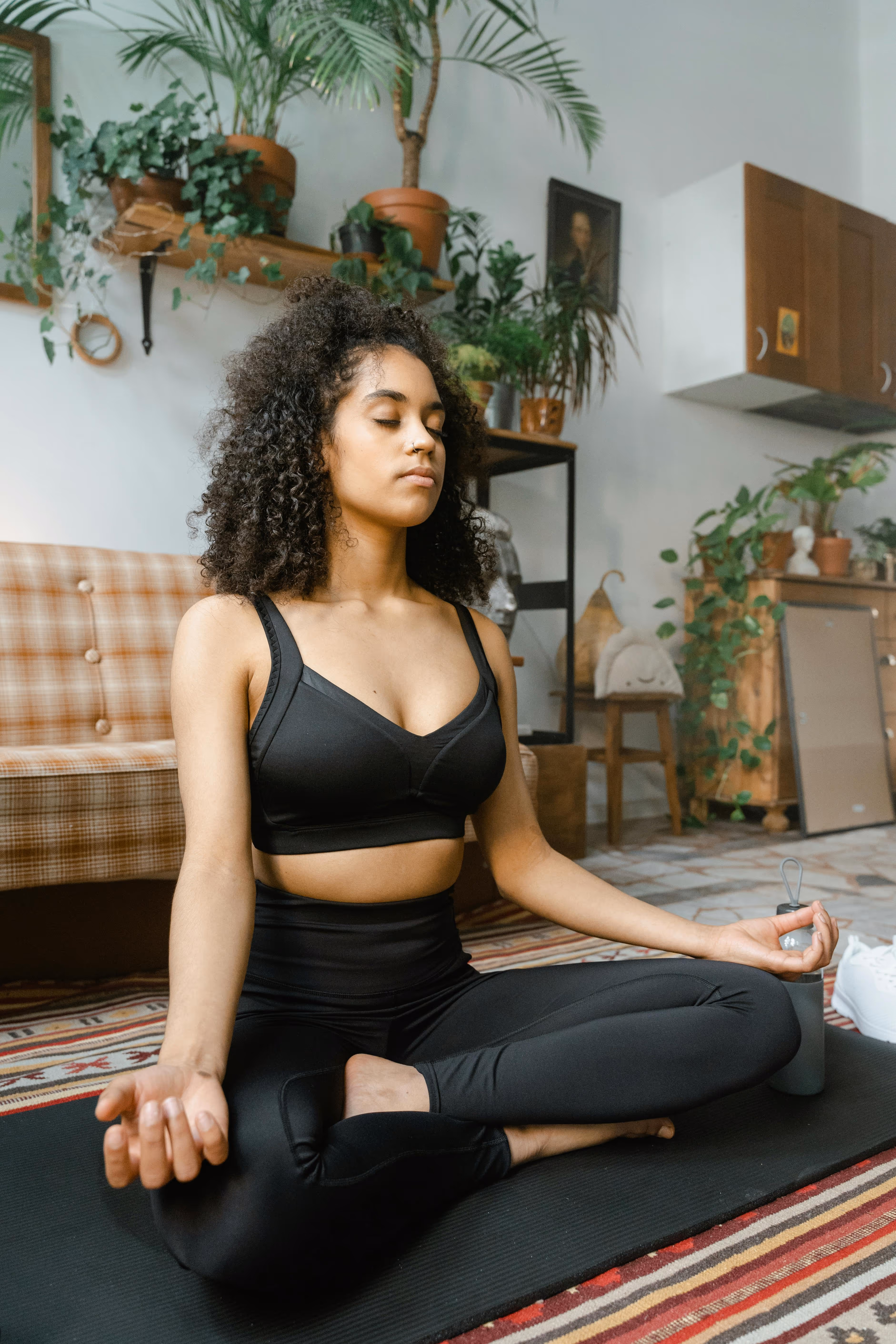 Young woman with curly hair wearing black workout clothes meditating cross-legged on a yoga mat indoors.