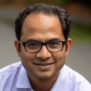 Smiling man wearing black glasses and a light purple shirt with a blurred outdoor background.
