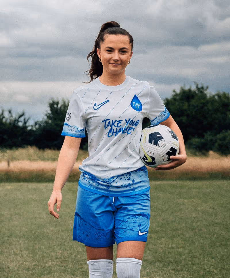 Smiling girl in a white and blue football kit that reads “Take Your Chance!” holding a football on a grassy pitch under cloudy skies.
