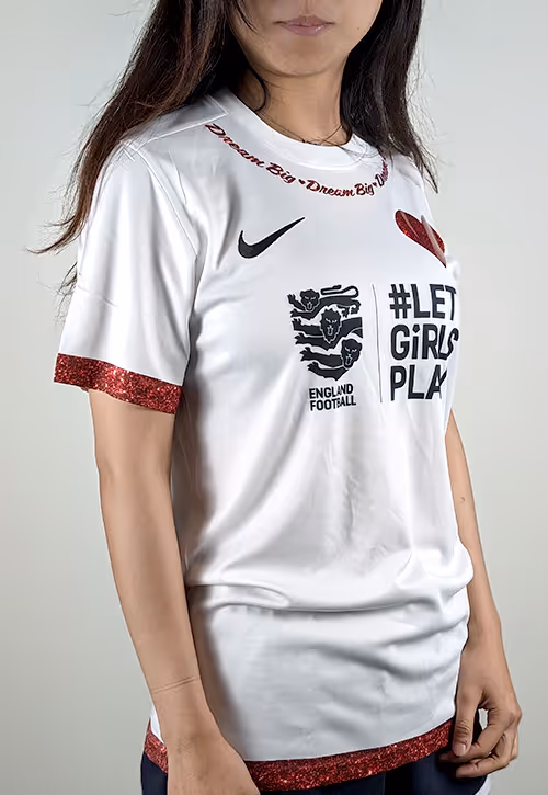Girl wearing a white football shirt with glittery red trim, a red heart patch, and bold black text reading “#LET GIRLS PLAY” alongside the England Football logo and Nike swoosh. “Dream Big” is written in cursive across the neckline.