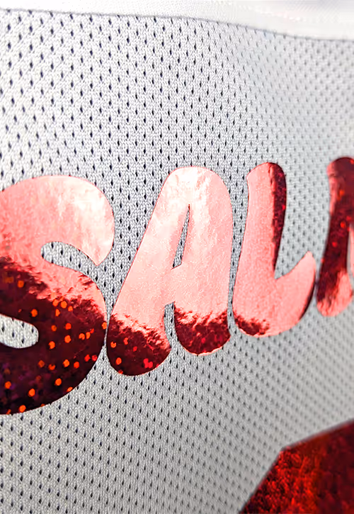 Close-up of a white football shirt with the name “SALMON” in shiny red glitter text, showing detailed texture on the mesh fabric and bold number partially visible.