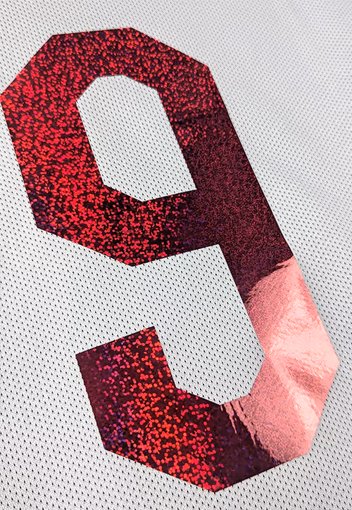 Close-up of a white football shirt with the numer 9 in shiny red glitter text, showing detailed texture on the mesh fabric and bold number partially visible.