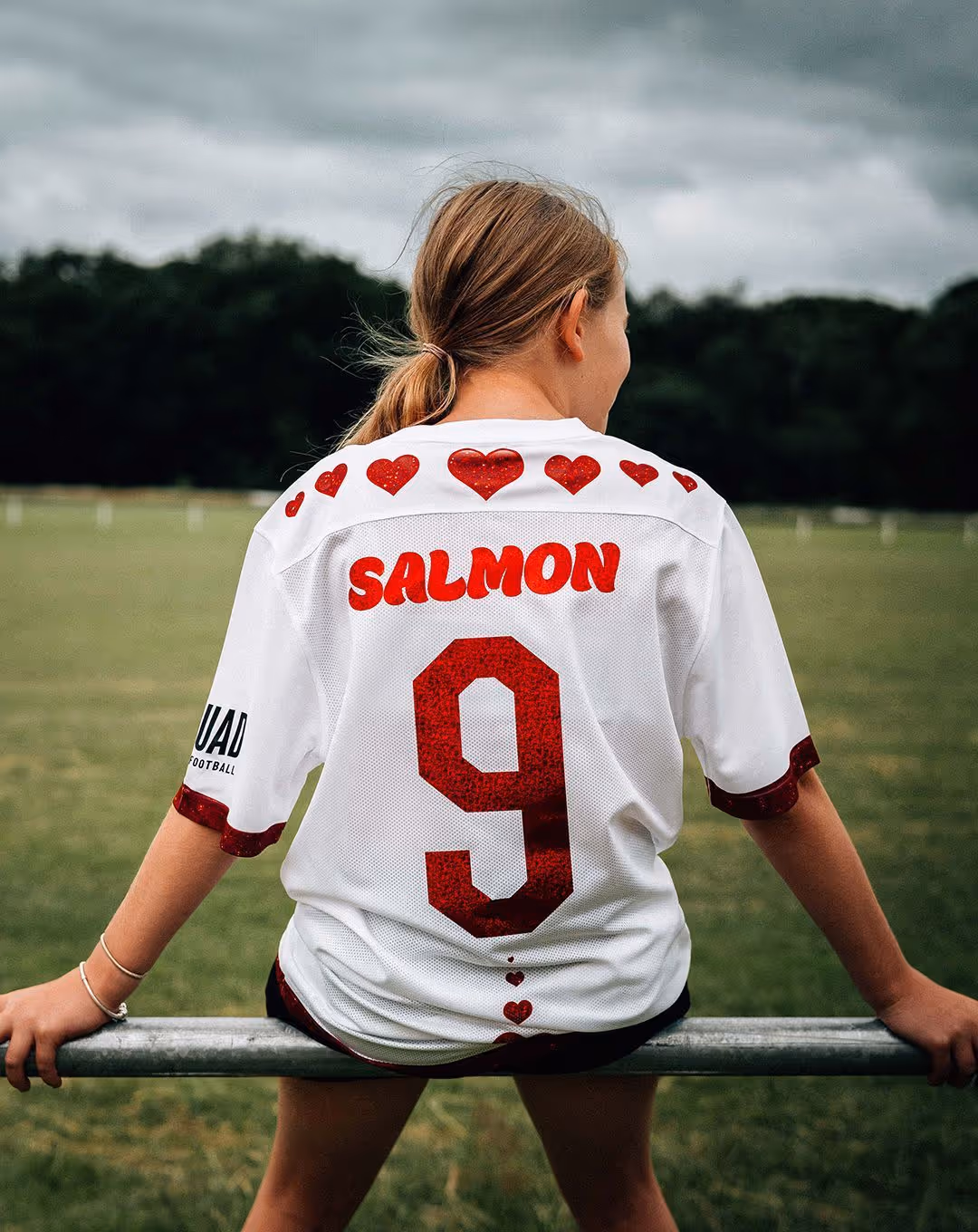 Girl seen from behind wearing a white football shirt with glittery red hearts, the name “SALMON,” and the number 9, leaning on a rail with a grassy pitch and trees in the background.