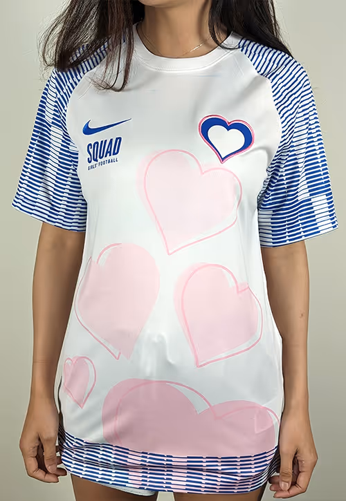 Girl wearing a white football shirt featuring blue striped sleeves, pink heart patterns, a stitched heart crest, and the text “SQUAD Girls’ Football” beside a blue Nike logo.