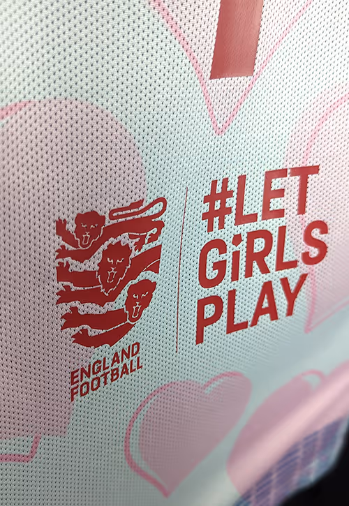 Close-up of a football shirt with the England Football logo and bold red text reading “#LET GIRLS PLAY,” surrounded by soft pink heart designs on a light background.
