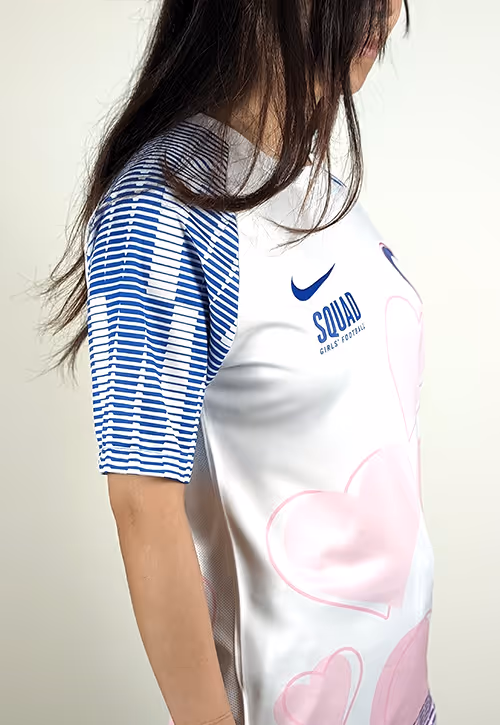 Girl wearing a white football shirt with bold pink heart graphics, blue striped sleeves, and the text “SQUAD Girls’ Football” beside a blue Nike logo.