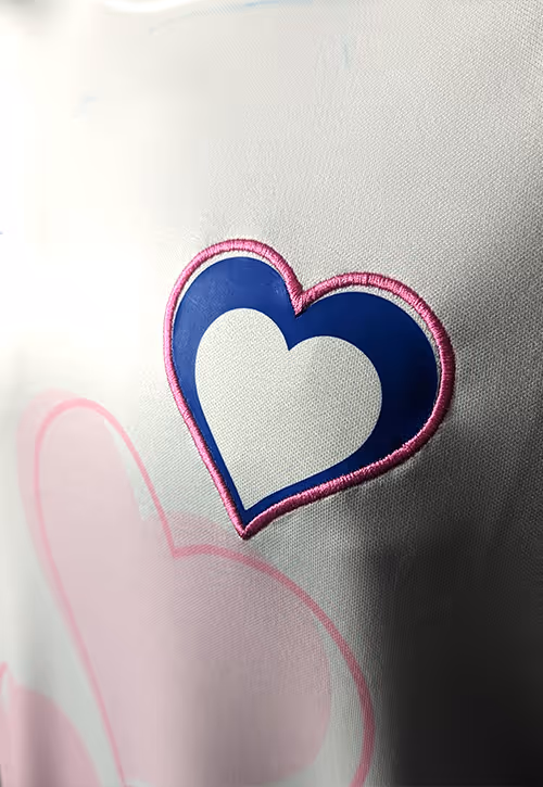 Close-up of a football shirt showing a stitched heart emblem in navy and white with a pink outline, layered over soft pink heart graphics on the fabric.