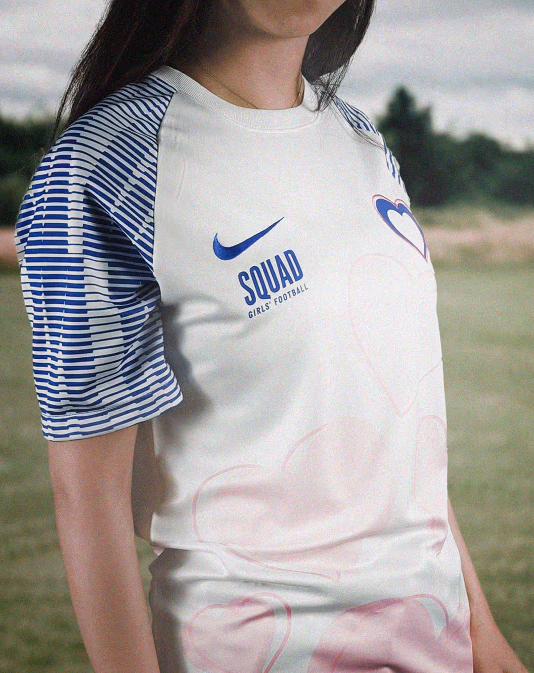 Close-up of a girl wearing a white football shirt with blue striped sleeves, pink heart patterns, and text reading “SQUAD Girls’ Football” and a blue Nike logo.
