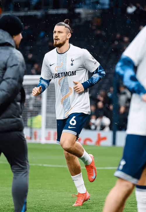 Footballer in a white Tottenham Hotspur kit with navy shorts and pink boots warming up on the pitch, with light rain falling and blurred players and staff in the background.