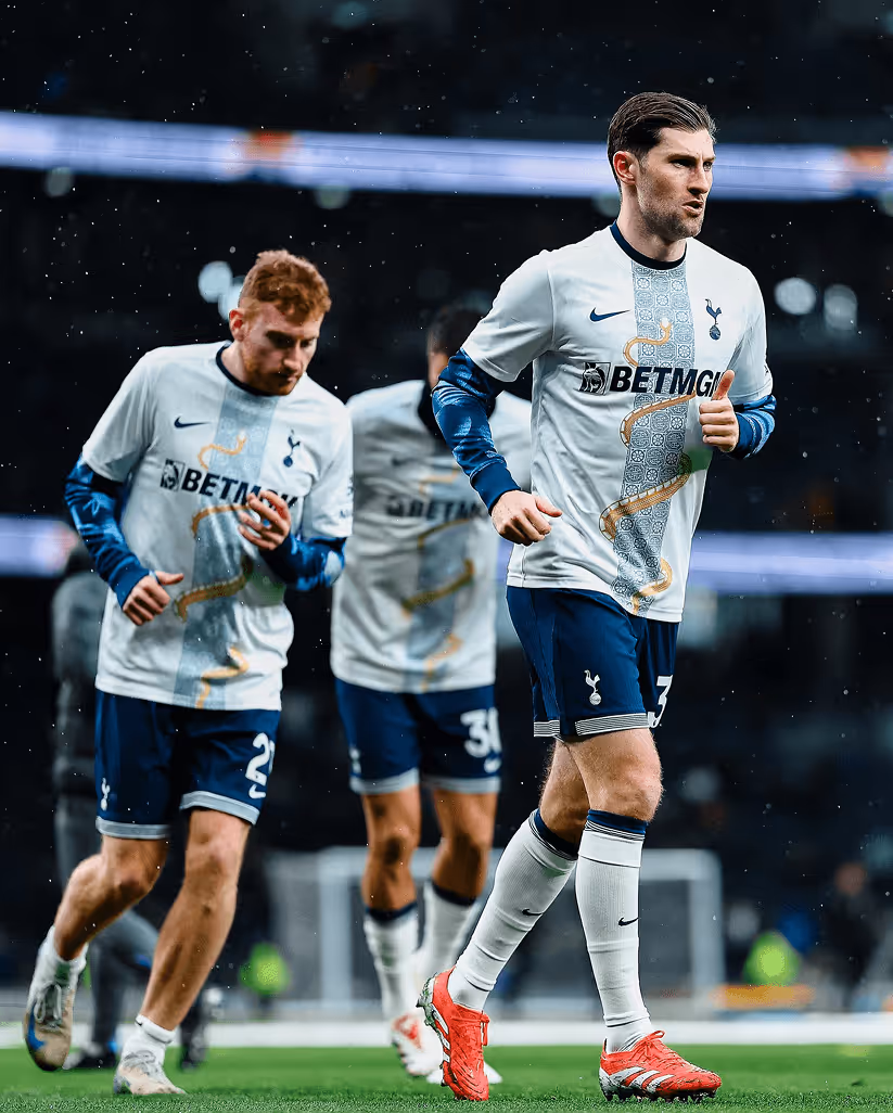 Tottenham Hotspur footballers warming up on the pitch in white and navy kits with a gold and blue design down the front, under light rain and stadium lights.