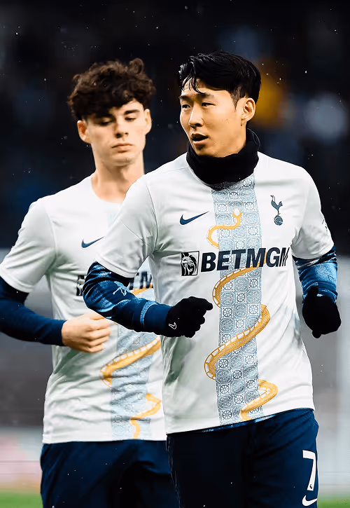 Tottenham Hotspur players warming up in white and navy kits with a gold serpent design; the player in front wears gloves and a neck warmer, with light rain falling around them.