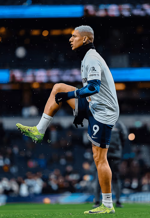 Tottenham Hotspur player stretching on the pitch in a white and navy kit with number 9 on his shorts, wearing bright green boots and gloves under light rain.