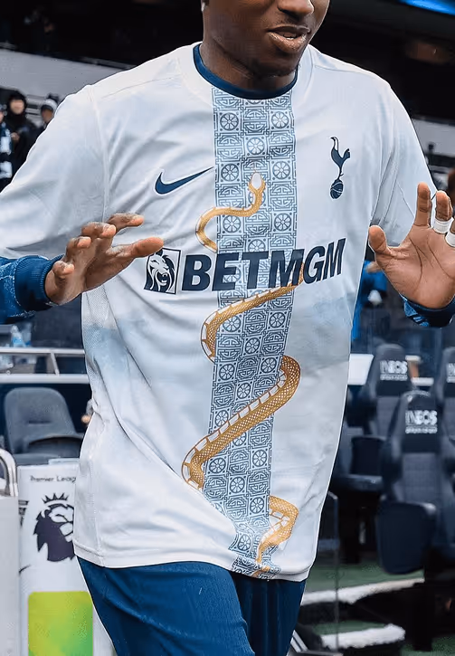 Tottenham Hotspur player smiling and walking past the dugout in a white and navy kit with a gold serpent design, with fans and stadium seating in the background.