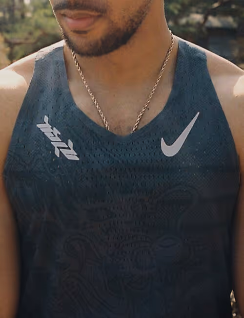 Close-up of a man wearing a dark athletic tank top with a subtle dragon pattern, featuring two logos: a stylised script on the left and a Nike Swoosh on the right. He also wears a gold chain necklace.