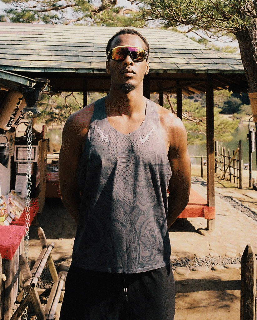 A man in a patterned athletic tank top and reflective sunglasses stands confidently outdoors in front of a traditional wooden structure, with trees and a lake in the background. The image has a subtle animated effect.