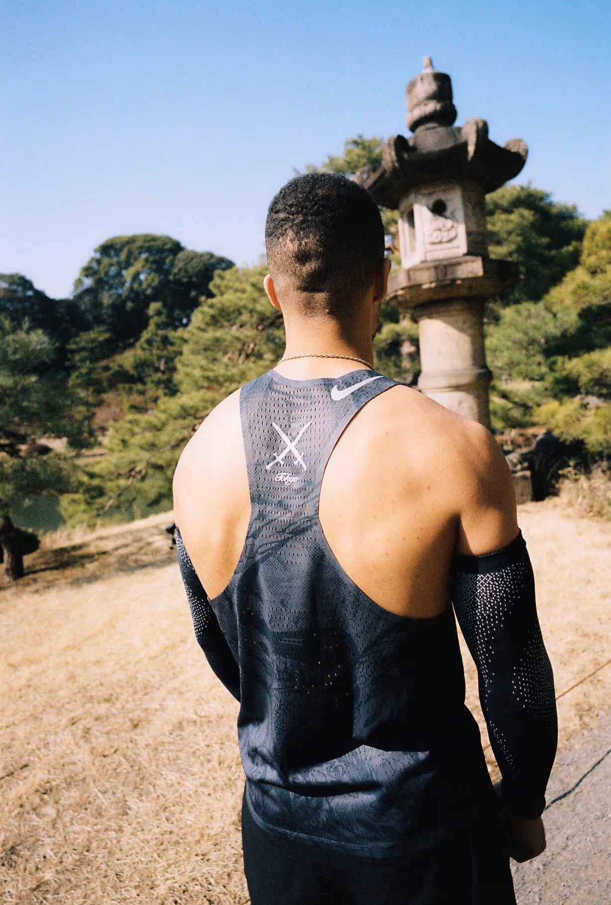 A man in a sleeveless athletic top stands with his back to the camera, facing a traditional Japanese stone lantern in a garden.