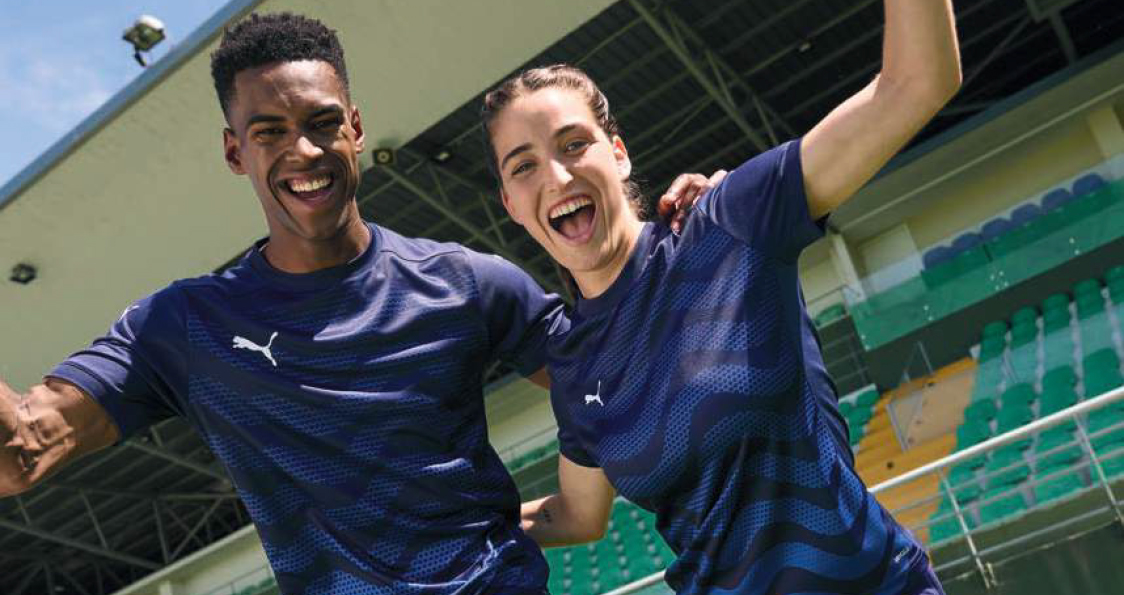 Two smiling athletes in matching navy blue Puma shirts pose with arms raised in a stadium, celebrating together in front of green and yellow seats.