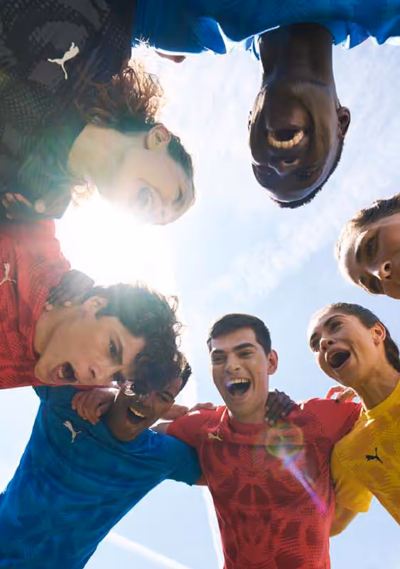 Group of young people in colorful Puma jerseys huddling and cheering under a bright sky, captured from below with sunlight streaming in.