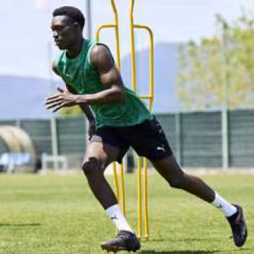 Male footballer in a green training bib and black shorts sprinting past a yellow mannequin on a grass field during a sunny training session.