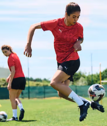 Female footballer in a red Puma jersey and black shorts juggling a ball mid-air on a sunny pitch, with another player practicing in the background.