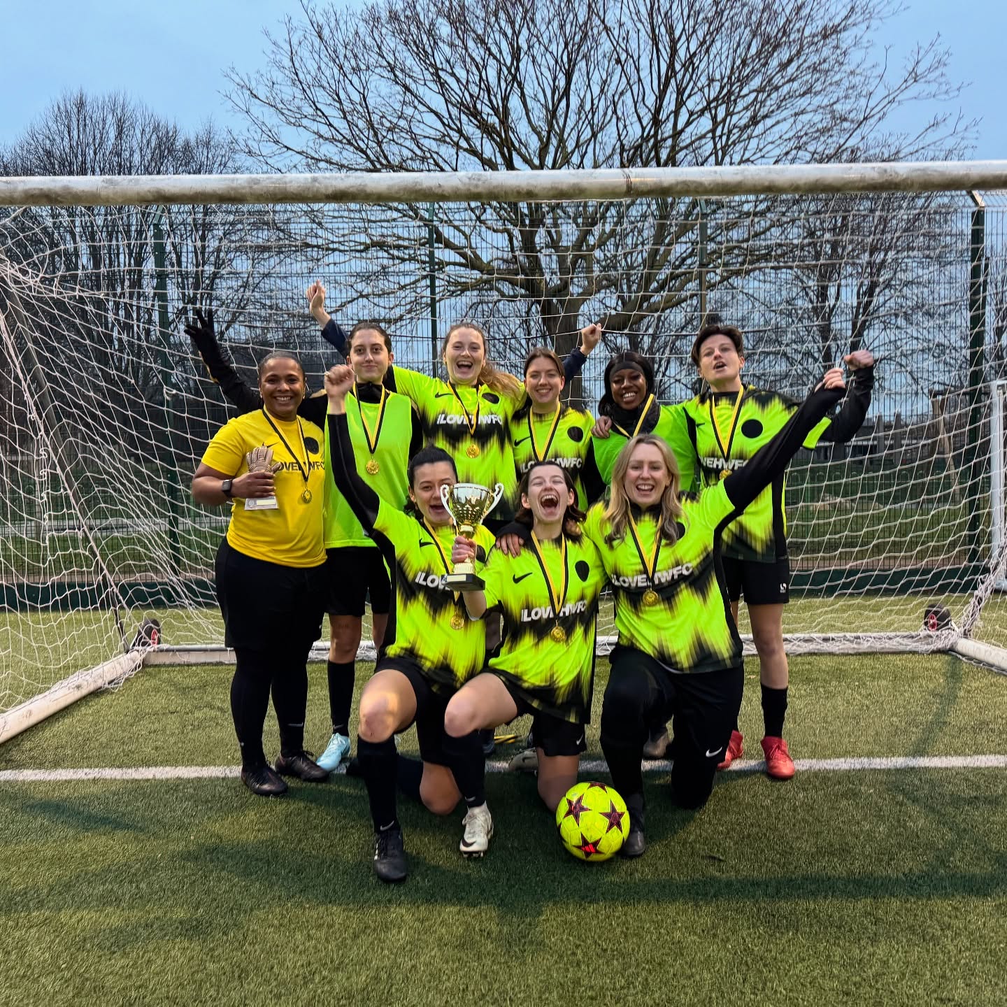 Female football team celebrating victory posing in the goal with the trophy and raised hands