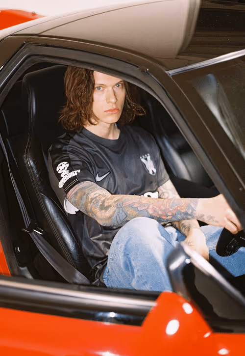 Young man with tattooed arms wearing a black sports jersey sitting in the driver's seat of a red car.
