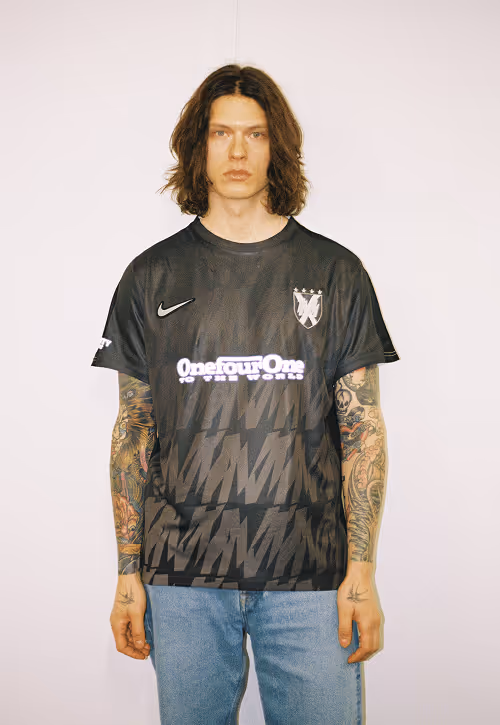 Male model with long hair standing against white wall wearing VESEY t-shirt and blue jeans