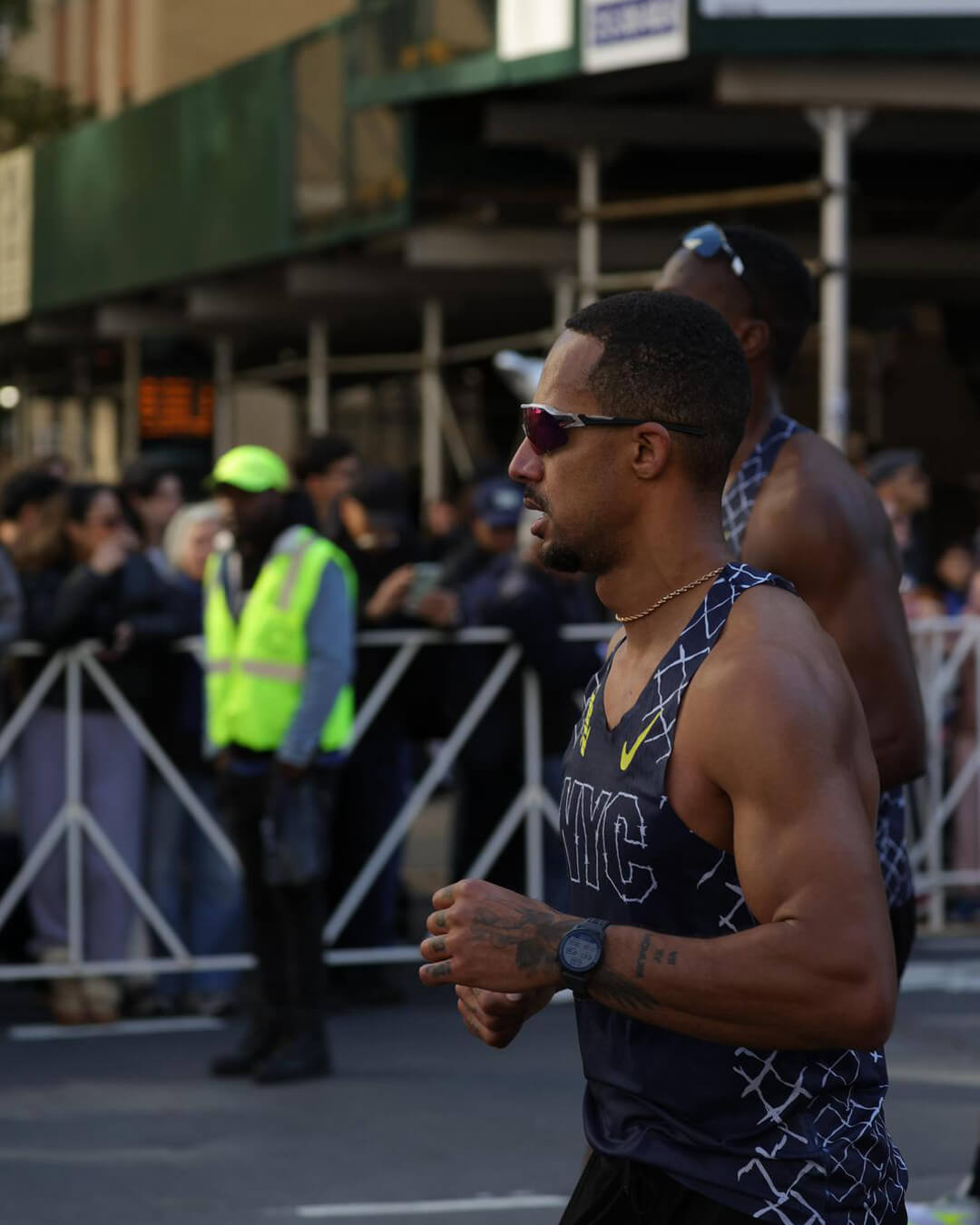 Runner in a NYC tank top and sunglasses participating in a city race with spectators and a race official in a neon vest in the background.