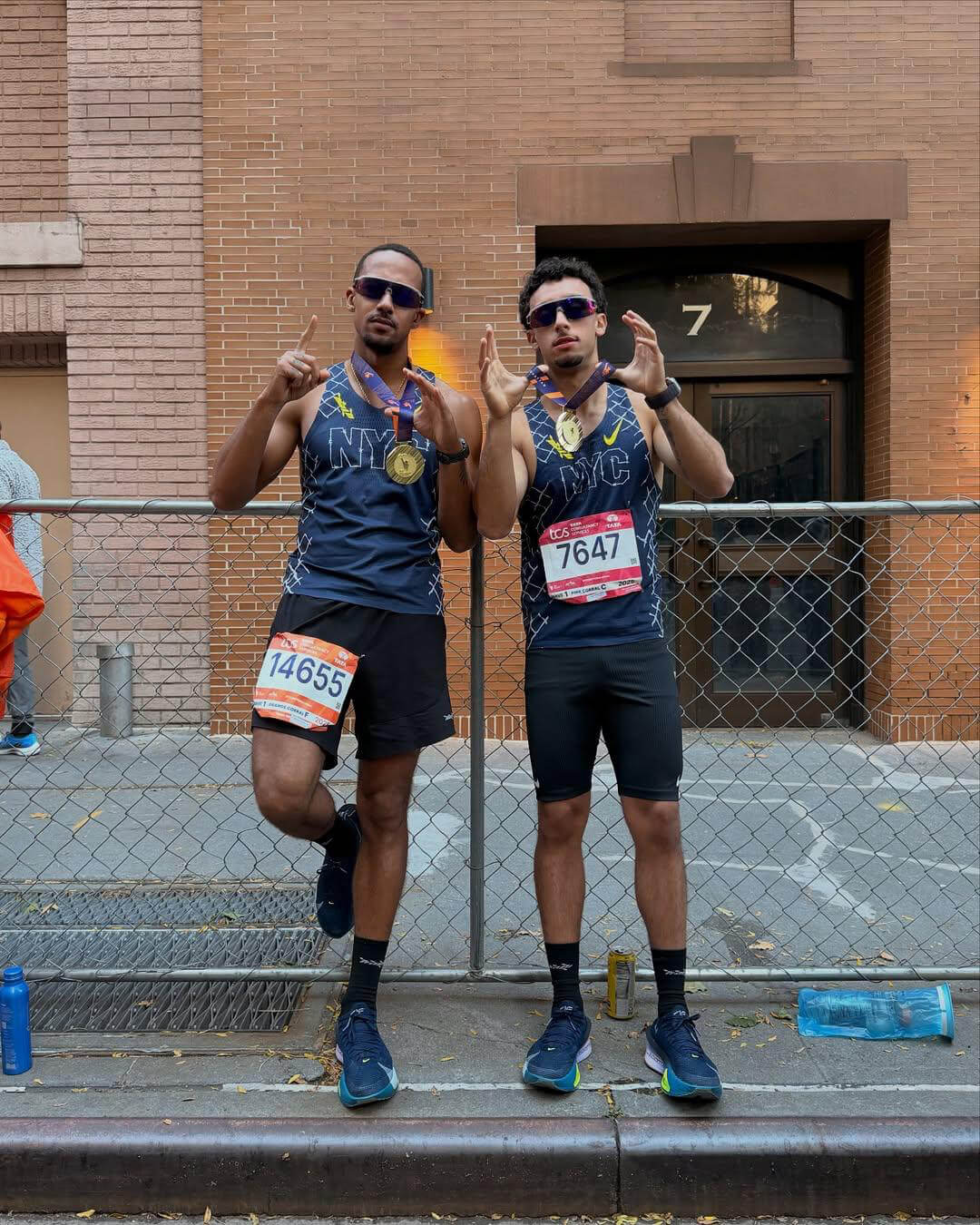 Two male runners wearing NYC race shirts and sunglasses, posing with gold medals and race bibs by a chain-link fence in front of a brick building.