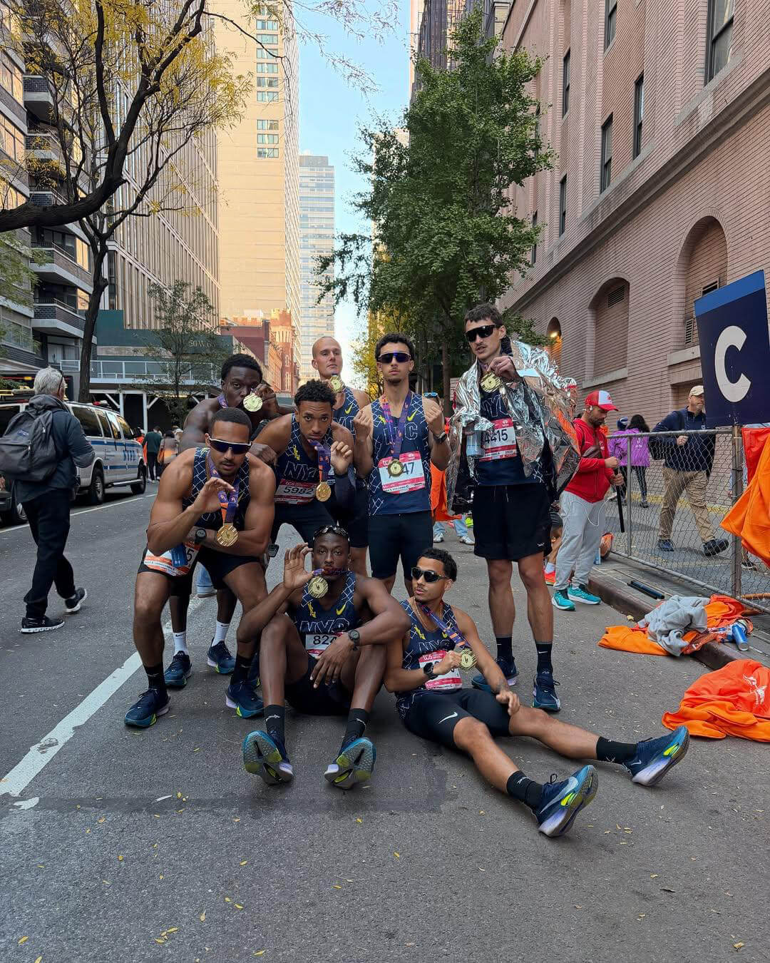 Seven male runners posing on a city street after a race, wearing medals and athletic gear, with tall buildings and trees in the background.
