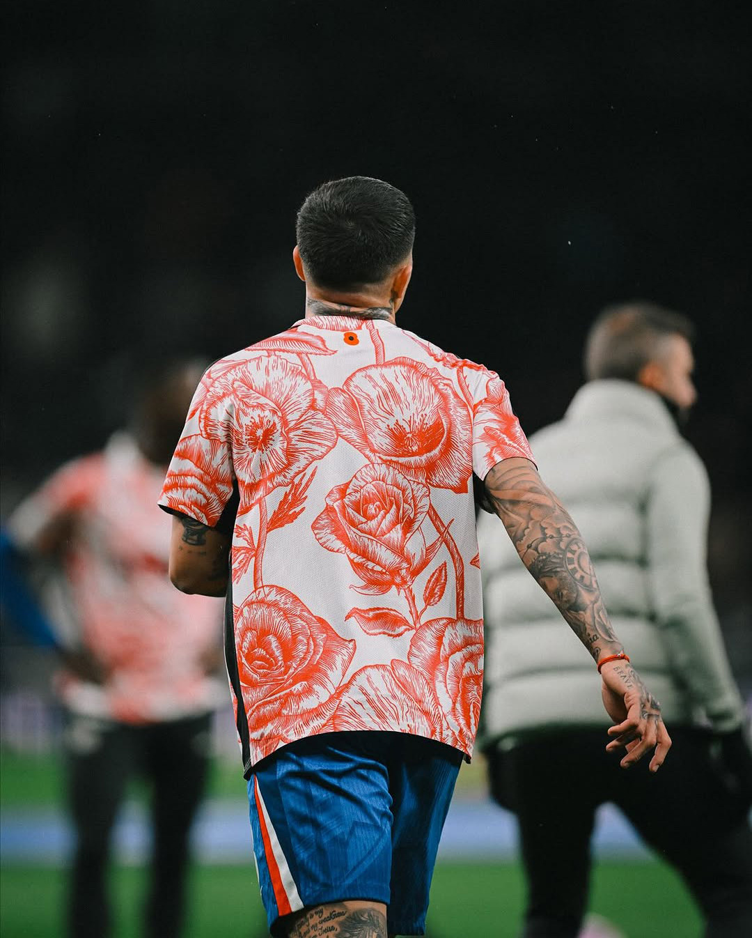 Man with tattoos wearing a white shirt with large red floral prints and blue shorts walking on a sports field.