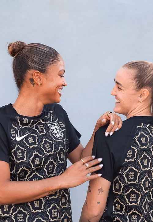 Two smiling women in black sports jerseys with gold patterns, facing each other against a gray background.