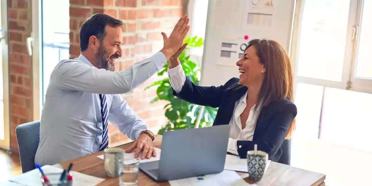 A man and woman shake hands in front of a laptop.