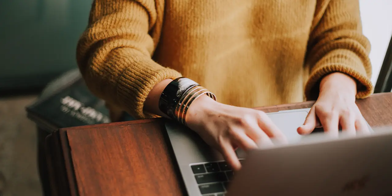 A woman wearing a yellow sweater and a watch is typing on a laptop.