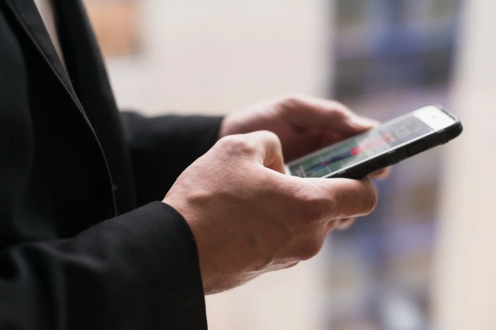 Primer plano de una persona vestida con un blazer negro usando un teléfono inteligente, con ambas manos sosteniendo el dispositivo y un fondo borroso en tonos suaves.