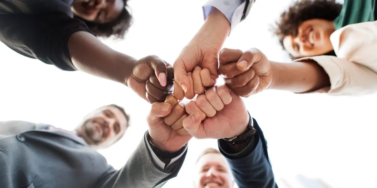 La imagen muestra a un grupo de personas en círculo, vistas desde abajo, juntando los puños en el centro en un gesto de unidad y trabajo en equipo. Las manos son de diferentes tonos de piel, lo que representa la diversidad. Todos sonríen, transmitiendo una atmósfera positiva de cooperación y asociación. Las personas visten ropa formal e informal, lo que sugiere un ambiente profesional inclusivo y amigable. El fondo es claro, con un cielo blanco en el fondo, que resalta el gesto colectivo.
