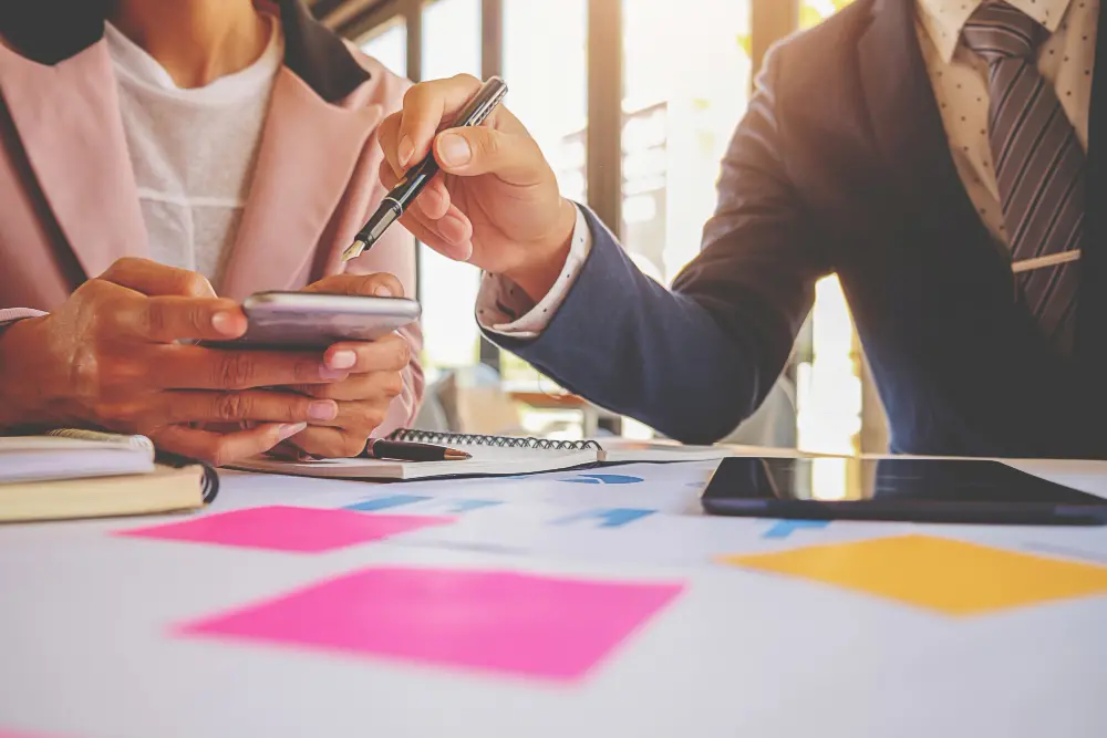 Duas pessoas analisando informações no smartphone durante uma reunião.