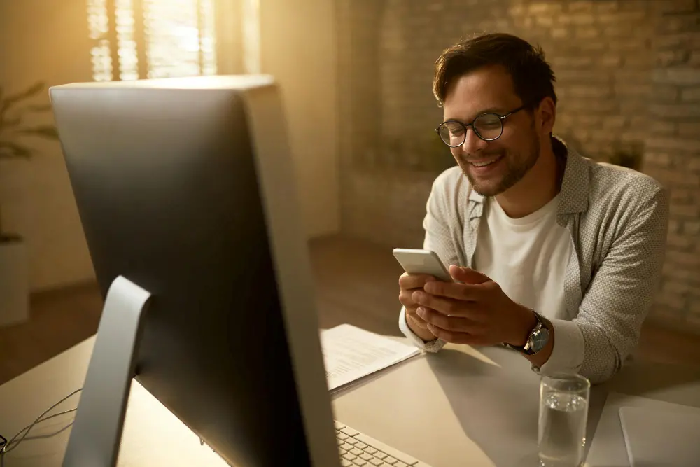 Man smiling while using a smartphone at a desk with a computer.