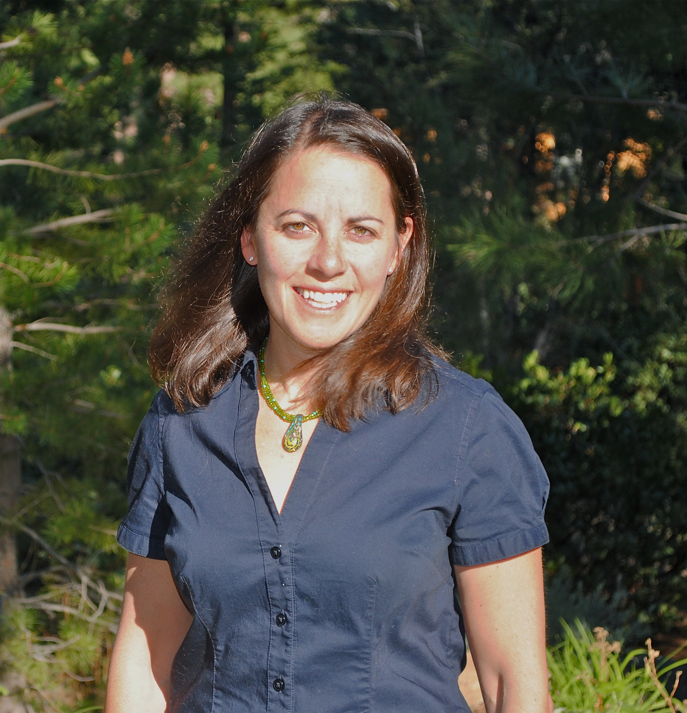 Smiling woman with shoulder-length brown hair wearing a navy blue button-up shirt and green pendant necklace standing outdoors with trees in the background.