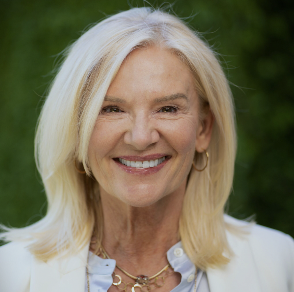 Smiling middle-aged woman with shoulder-length blonde hair, wearing gold hoop earrings, layered gold necklaces, a light blue blouse, and a white blazer, with a blurred green background.