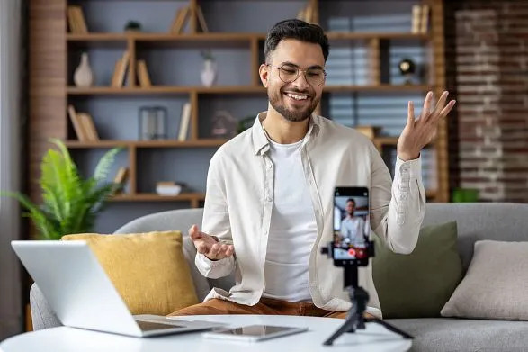 Smiling man recording a video on smartphone for social media, seated on couch with laptop, in a modern home office setup.