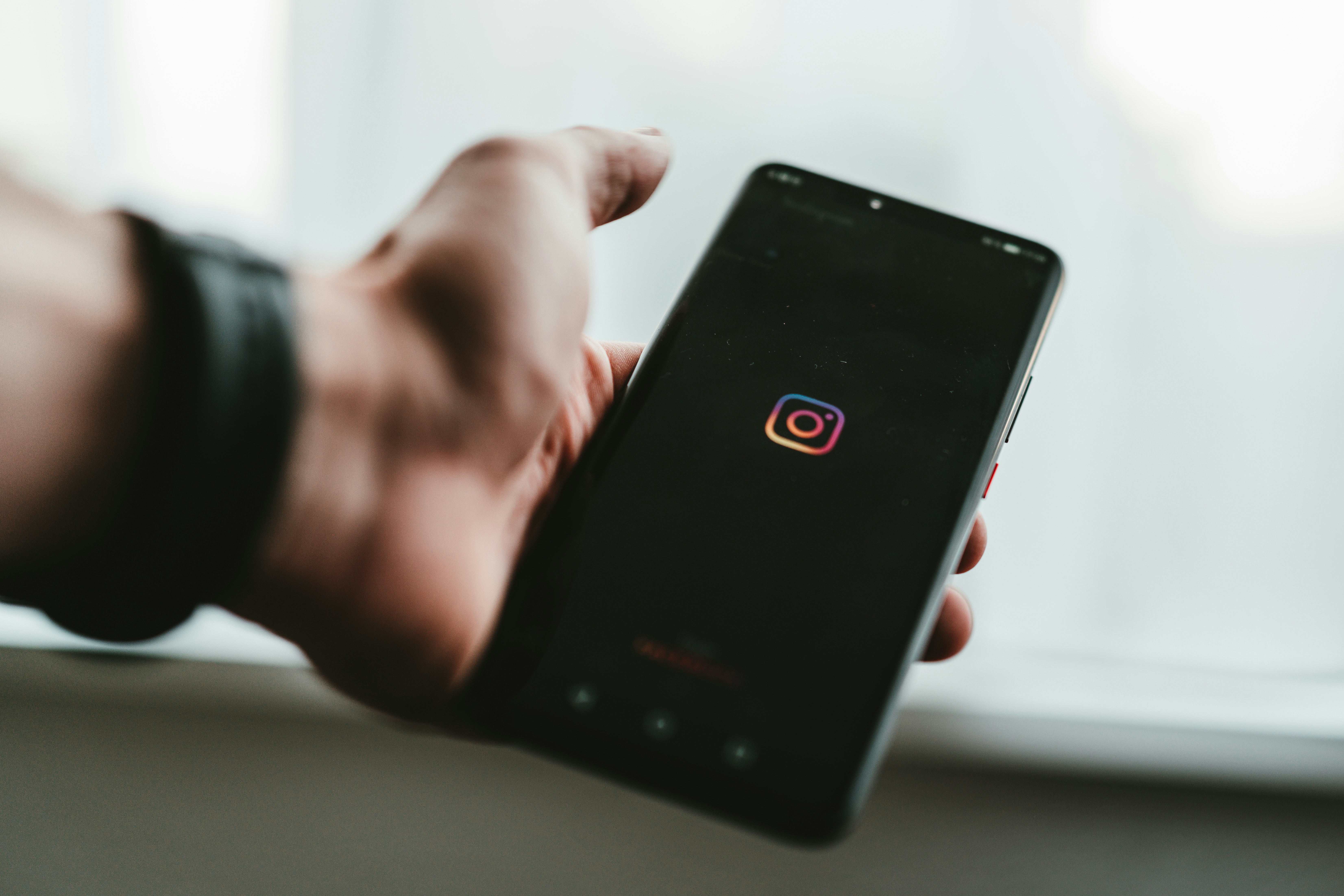 Hand holding a smartphone displaying the Instagram logo on a dark screen with a blurred white background. 