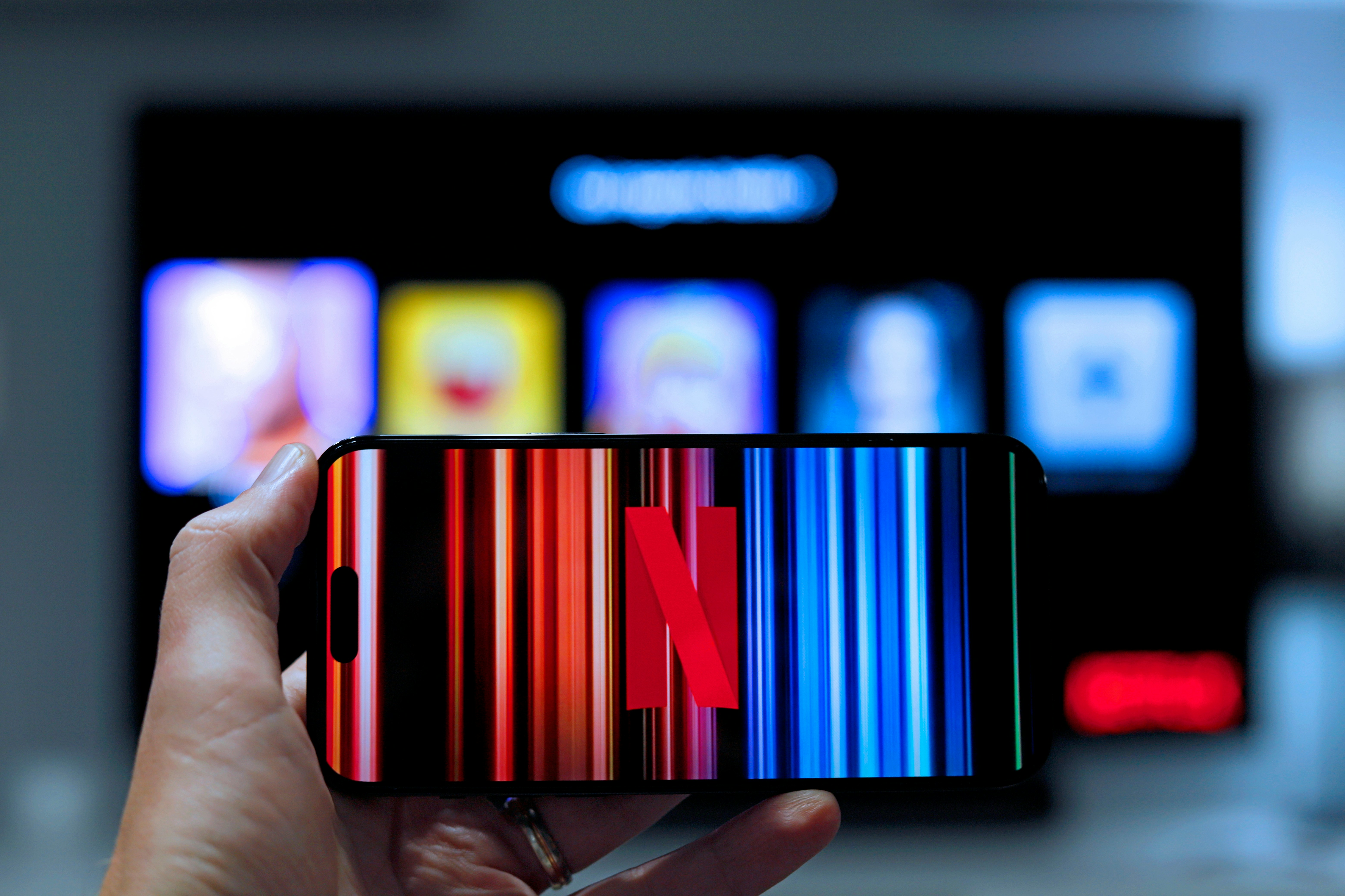 Hand holding smartphone with Netflix logo on vibrant vertical stripes, streaming profiles blurred on TV in modern living room background.