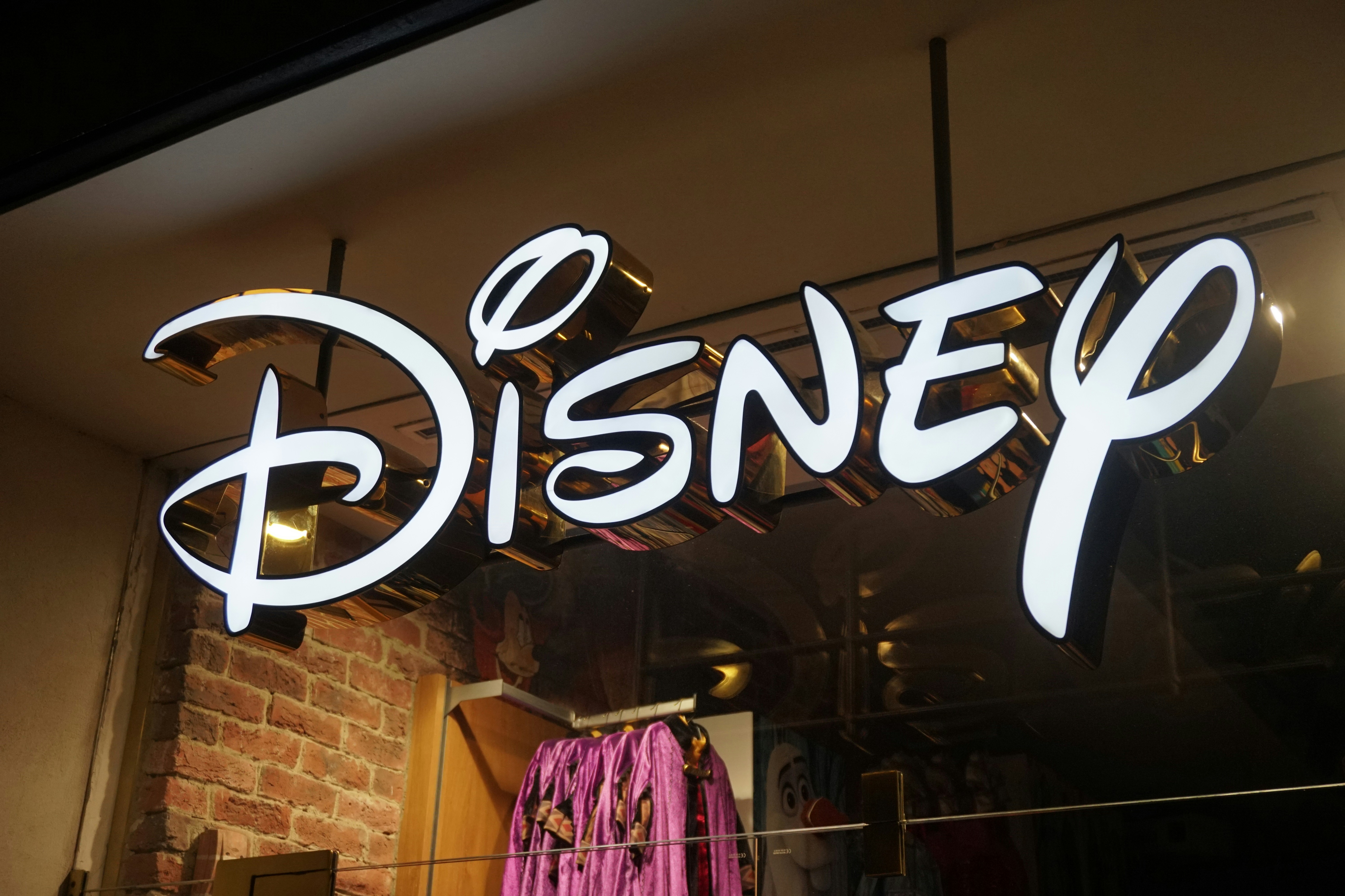 A brightly lit Disney logo sign hangs above a store entrance, featuring the iconic stylized script in white with gold edges. 