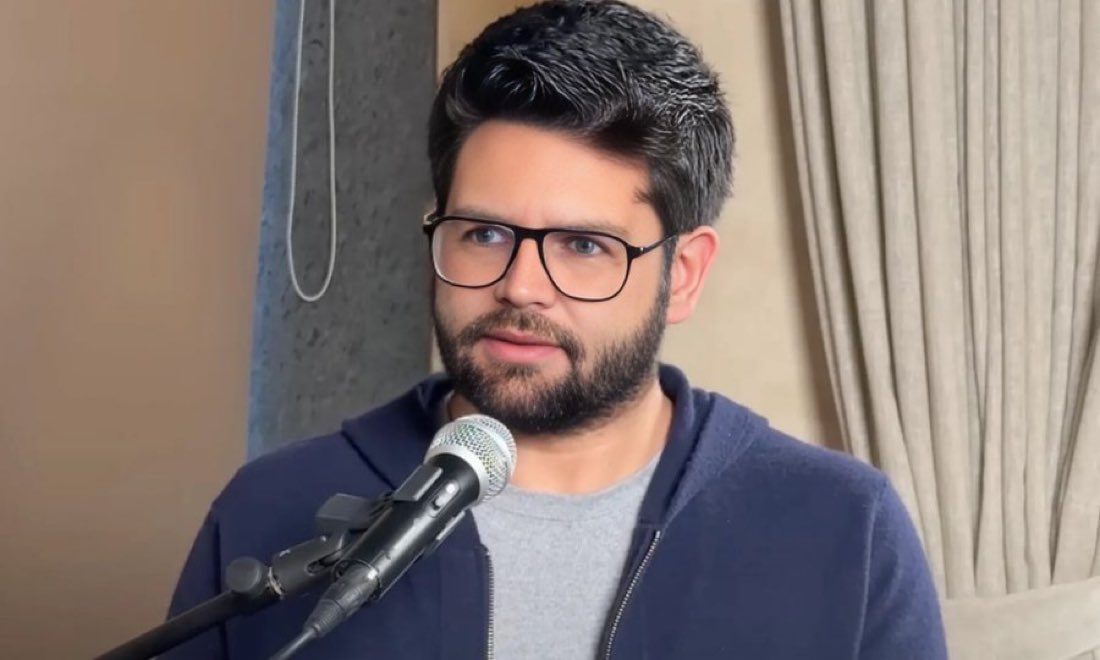 Perplexity CEO, Aravind Srinivas, speaking into a microphone, wearing a navy hoodie and gray shirt indoors.