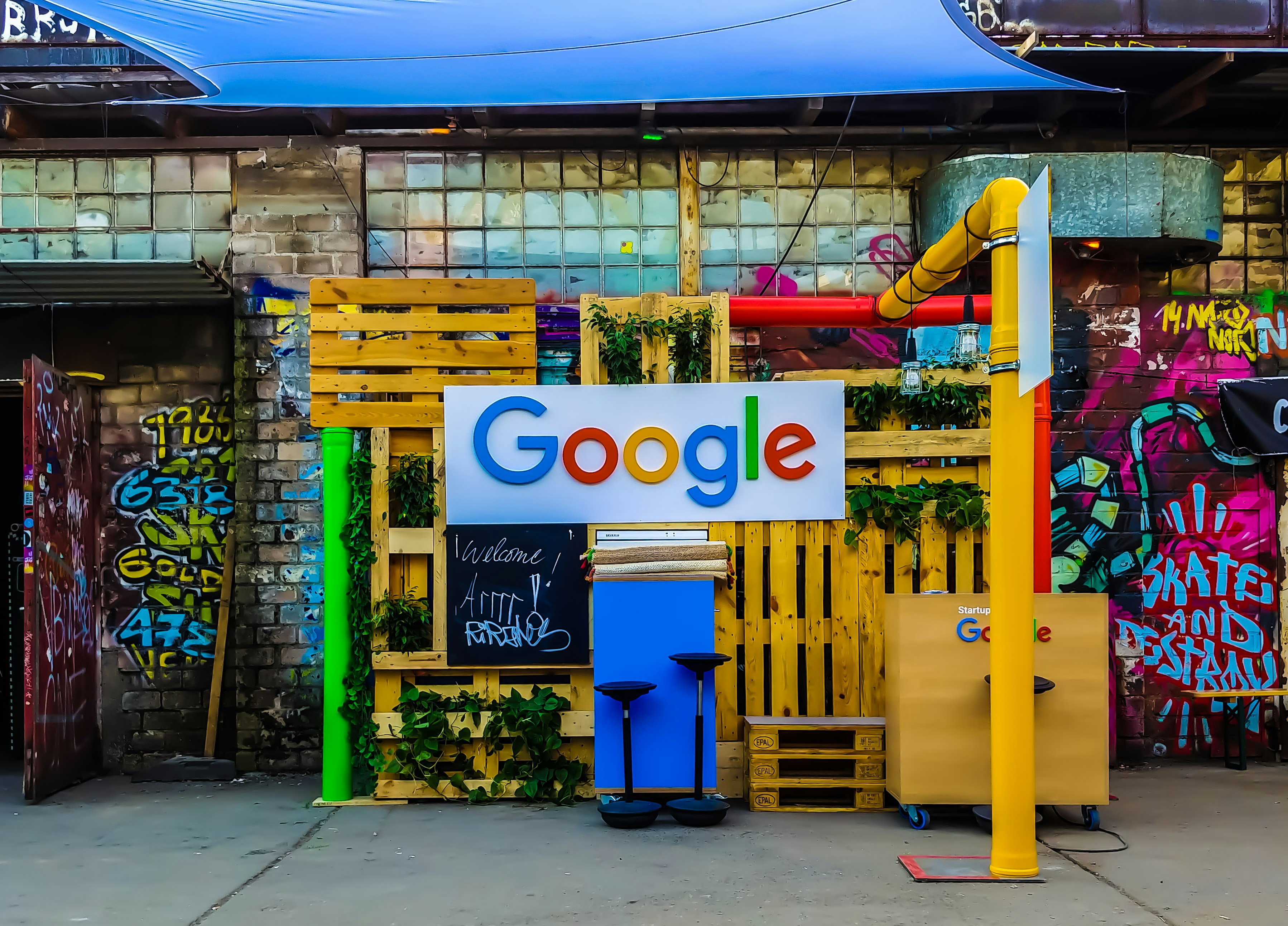 Colorful Google logo on a pallet wall with plants, graffiti, and a startup setup in a vibrant urban space.