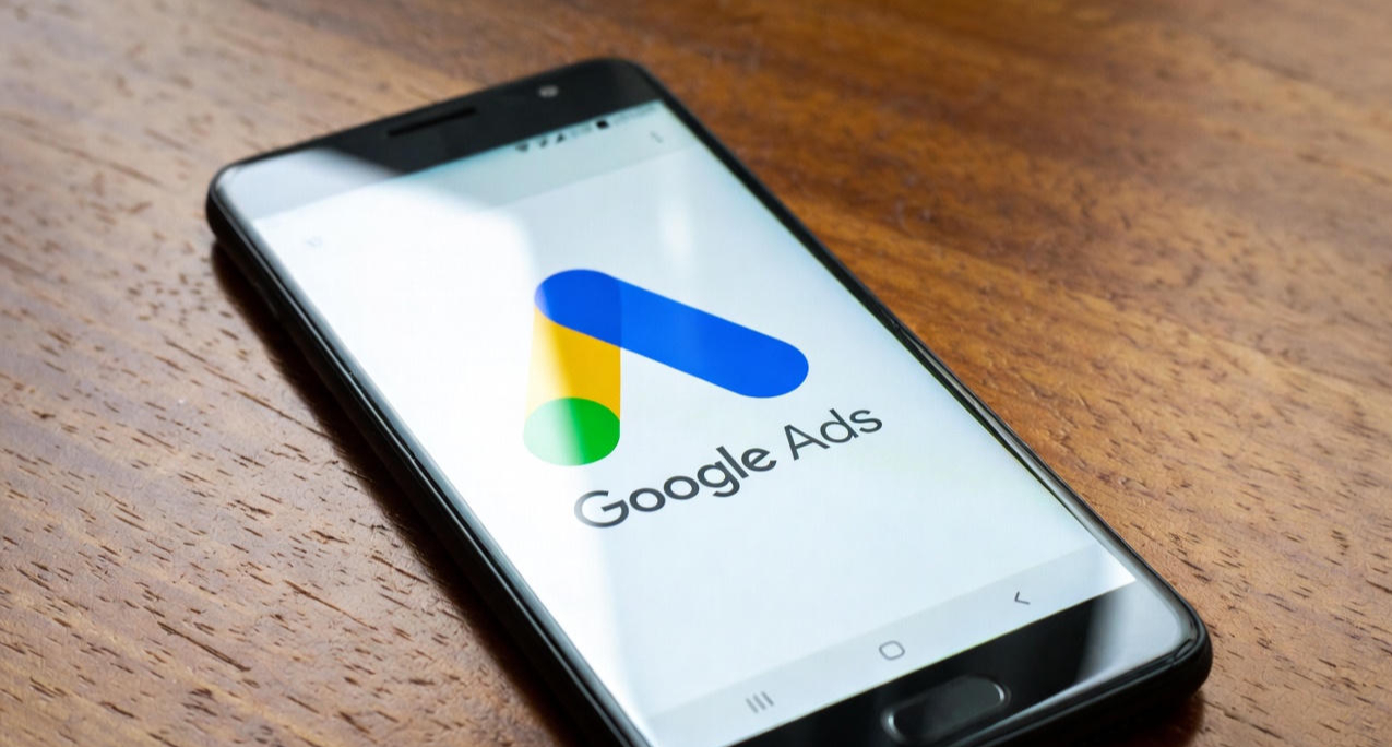  Smartphone tilted left on a wooden surface displaying a colorful Google Ads logo on the screen with realistic reflections.