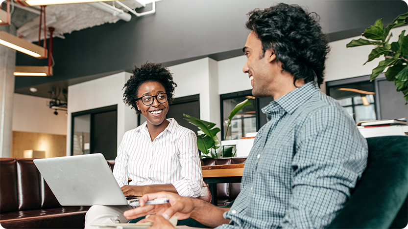  Two colleagues smiling and collaborating on a laptop in a modern coworking space with exposed ceilings and plants.